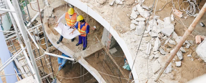 Construction Workers Looking Over Blue Prints In Broken Building Structure.