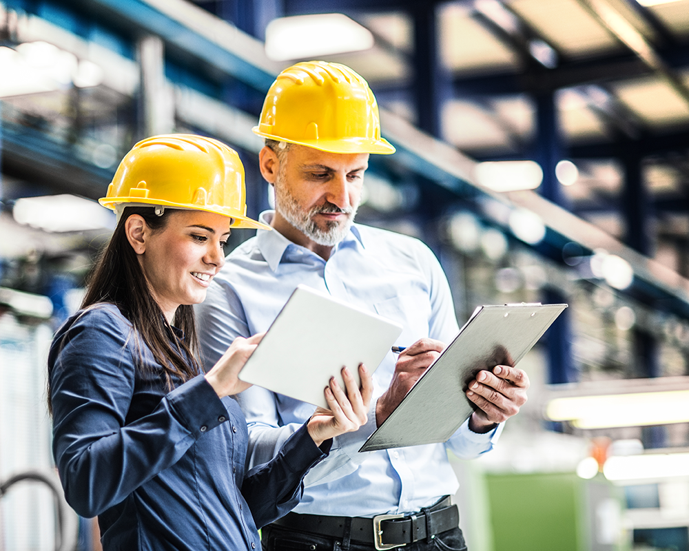 Two construction workers talking while looking at a tablet and clipboard.