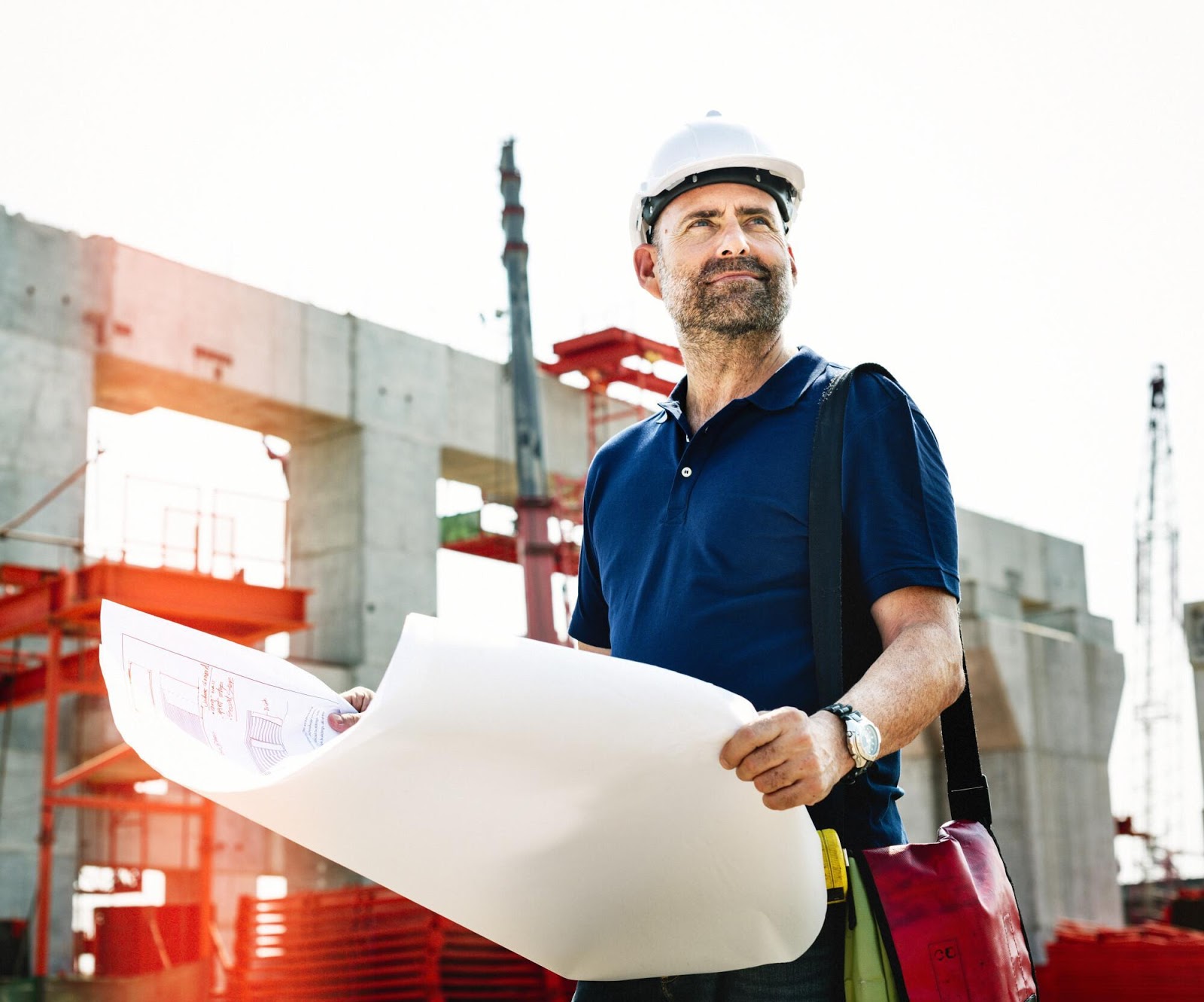 man construction worker holding blueprints looking in the distance at a construction site.