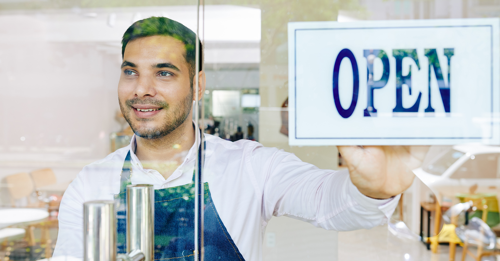 Man wearing an apron looking through the window while touching the Open sign.
