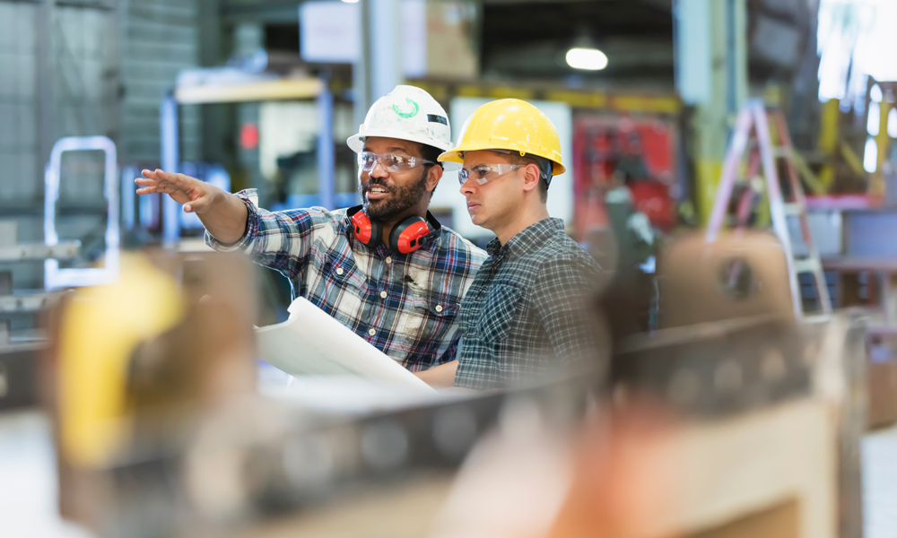 two male construction workers talking while one holds a blueprint and the other points out in the view.