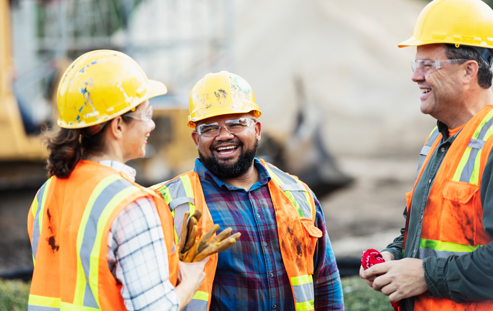 Three construction workers smiling and chatting in a circle on a construction site.