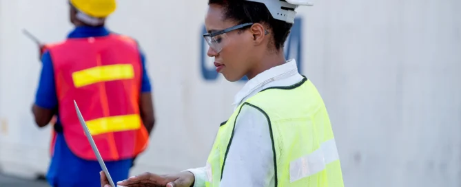 African American Woman In Construction Uniform Typing On Her Computer With African American Man In The Distance At Construction Site.