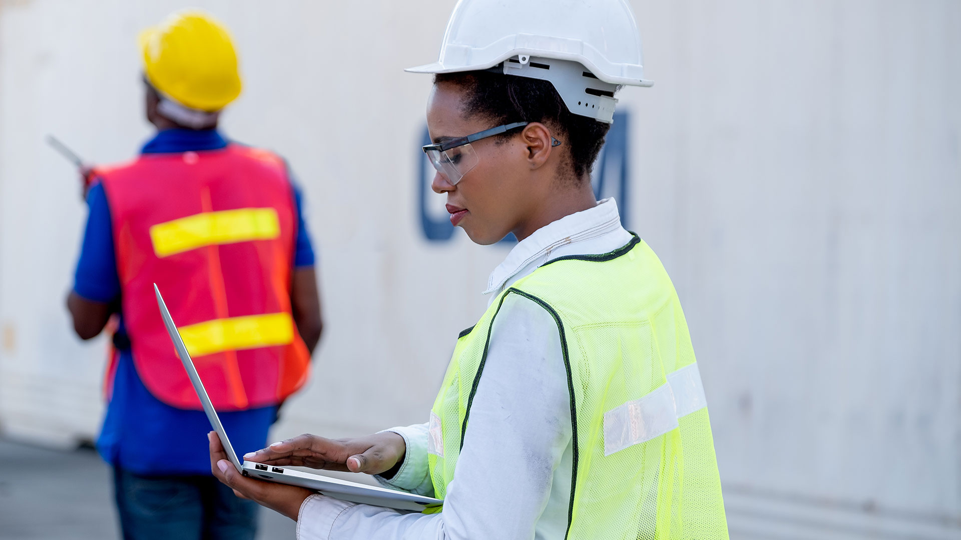 African American woman in construction uniform typing on her computer with African American man in the distance at construction site.